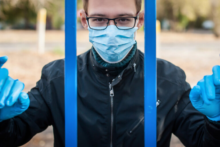A Man In A Medical Mask And Gloves Behind A Metal Bars In The St