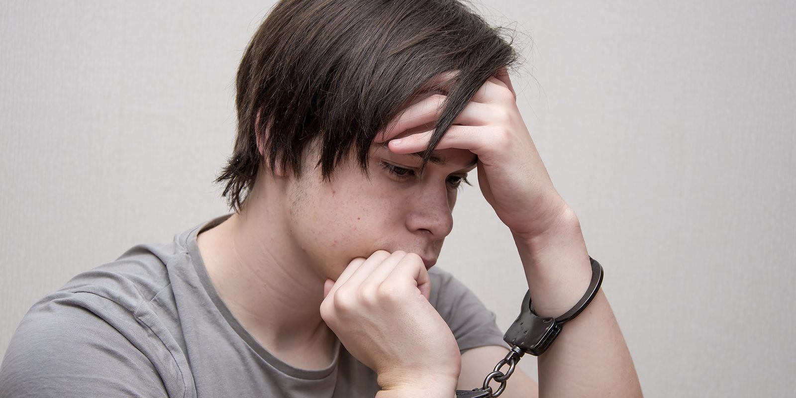 A Handcuffed Teenager Sits On A Grey Background. Juvenile Delinq