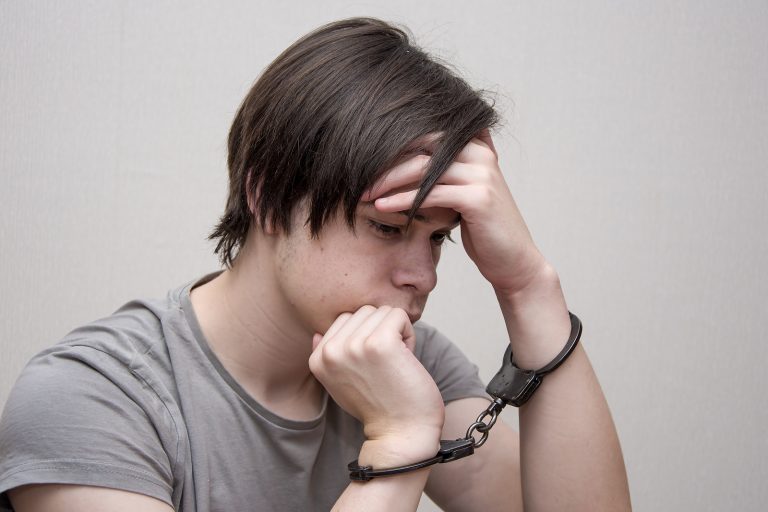 A Handcuffed Teenager Sits On A Grey Background. Juvenile Delinq
